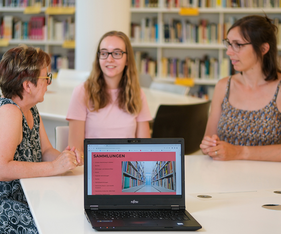 Drei Personen unterhalten sich, im Hintergrund ein Bücherregal, im Vordergrund ein Notebook, auf dem die Seite "Sammlungen" auf der Website der Deutschen Nationalbibliothek geöffnet ist. 