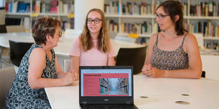 Drei Personen unterhalten sich, im Hintergrund ein Bücherregal, im Vordergrund ein Notebook, auf dem die Seite "Sammlungen" auf der Website der Deutschen Nationalbibliothek geöffnet ist. 