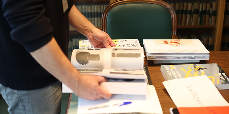 Eine Person blättert in einem Buch. Rund herum liegen weitere Bücher auf einem Tisch. Zu sehen ist außerdem ein Bücherregal und ein grüner Stuhl. Eine Person blättert in einem Buch. Rund herum liegen weitere Bücher auf einem Tisch. Zu sehen ist außerdem ein Bücherregal und ein grüner Stuhl.