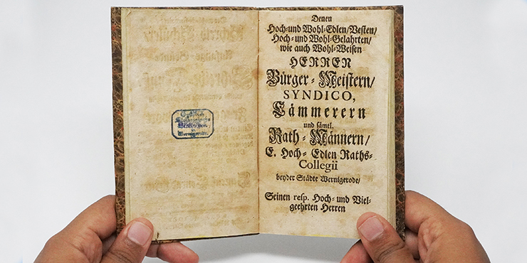 Zwei Hände halten ein Buch in der Hand. Es ist geöffnet und es ist ein Stempel auf linken sowie gedruckte Schrift auf der rechten Seite zu sehen. Zwei Hände halten ein Buch in der Hand. Es ist geöffnet und es ist ein Stempel auf linken sowie gedruckte Schrift auf der rechten Seite zu sehen.