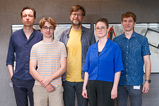 Personen sind für ein Gruppenfoto vor einer grau melierten Wand aufgestellt. Personen sind für ein Gruppenfoto vor einer grau melierten Wand aufgestellt.