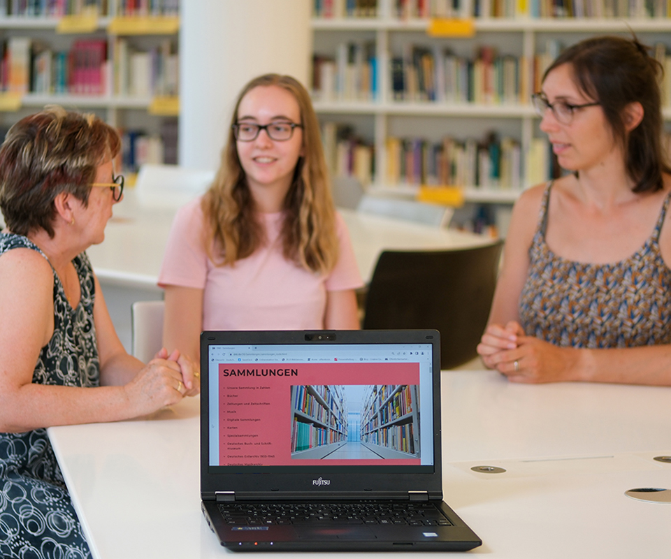 Three people talking, a bookshelf in the background, a notebook in the foreground, on which the “Collections“ page on the website of the German National Library is opened. Three people talking, a bookshelf in the background, a notebook in the foreground, on which the “Collections“ page on the website of the German National Library is opened.